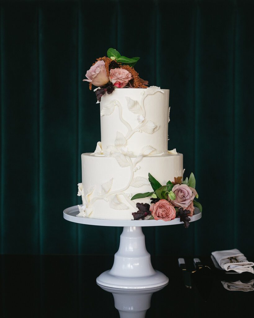 A two-tier white wedding cake with textured leaf detailing and fresh floral accents, set against an emerald velvet backdrop at an MDRD Grand Plaza wedding.