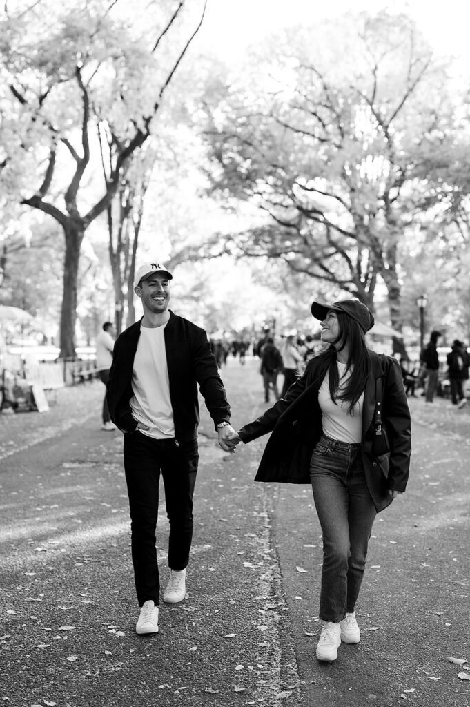 Black and white photo of a couple holding hands and laughing as they walk through Central Park.