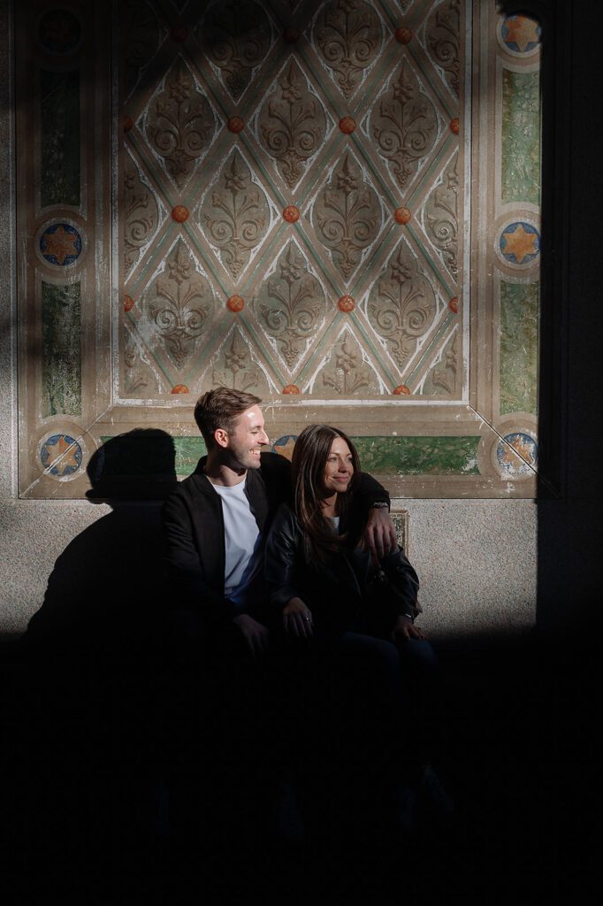 Couple sitting together in the shadows of Bethesda Terrace Arcade with patterned walls in the background.