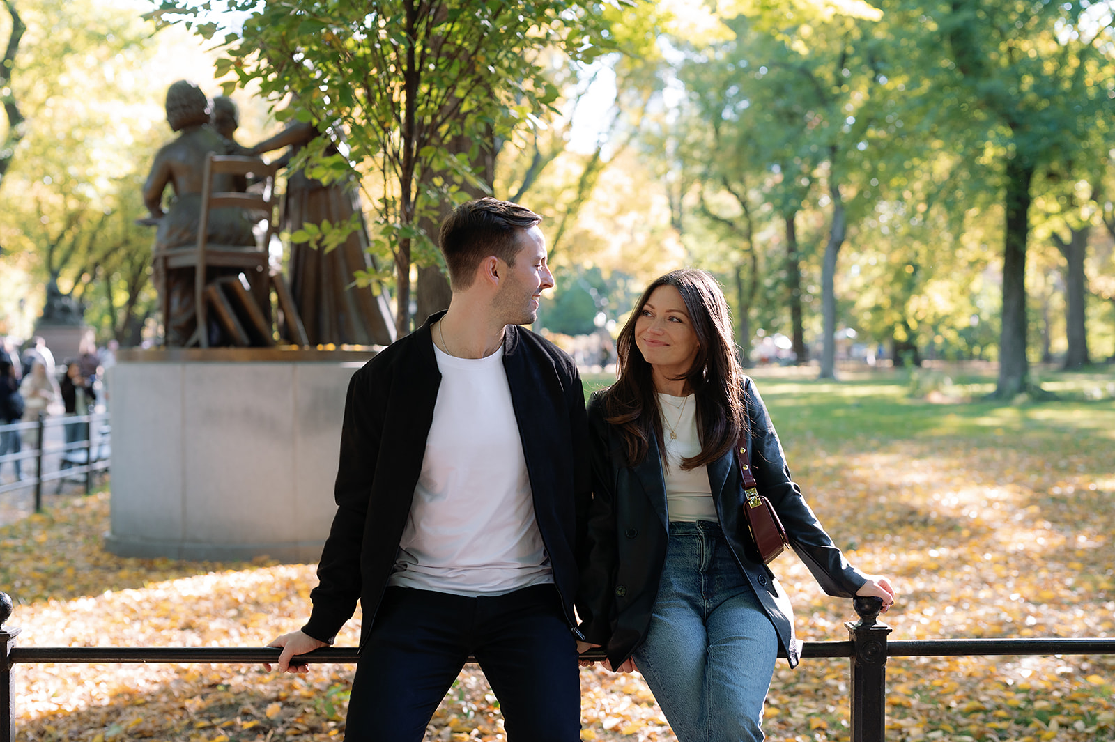 Couple smiling at each other while sitting near the Literary Walk statues in Central Park with autumn leaves on the ground.