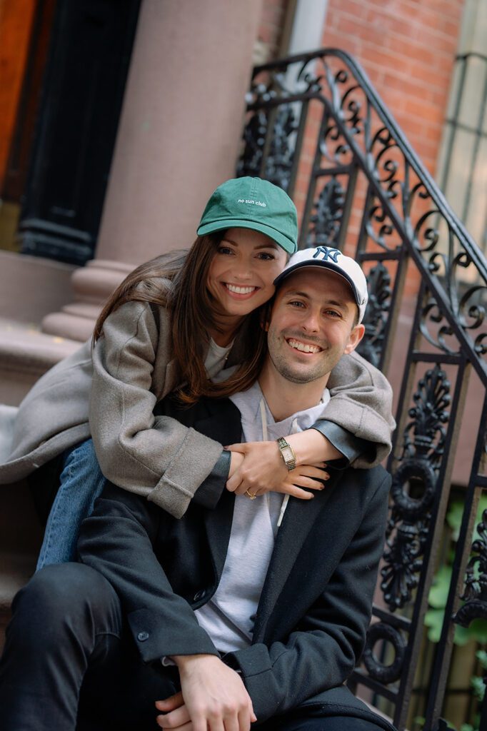 Color photo of a couple sitting on brownstone steps, the woman wrapping her arms around the man from behind during their Greenwich Village engagement photos.
