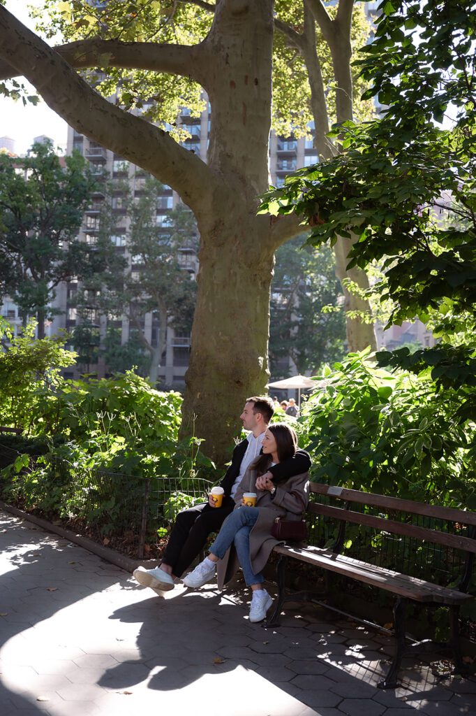 Couple sitting on a park bench with coffee cups, surrounded by trees, creating a cozy fall NYC engagement photo.