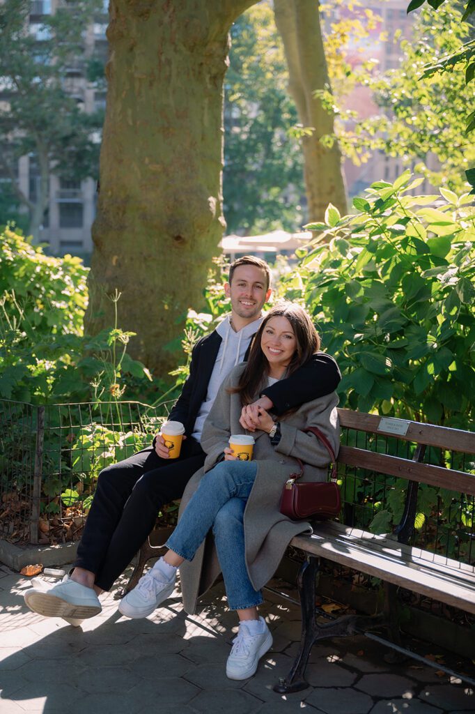 Couple smiling and sitting on a park bench with coffee cups, surrounded by trees, creating a cozy fall NYC engagement photo.