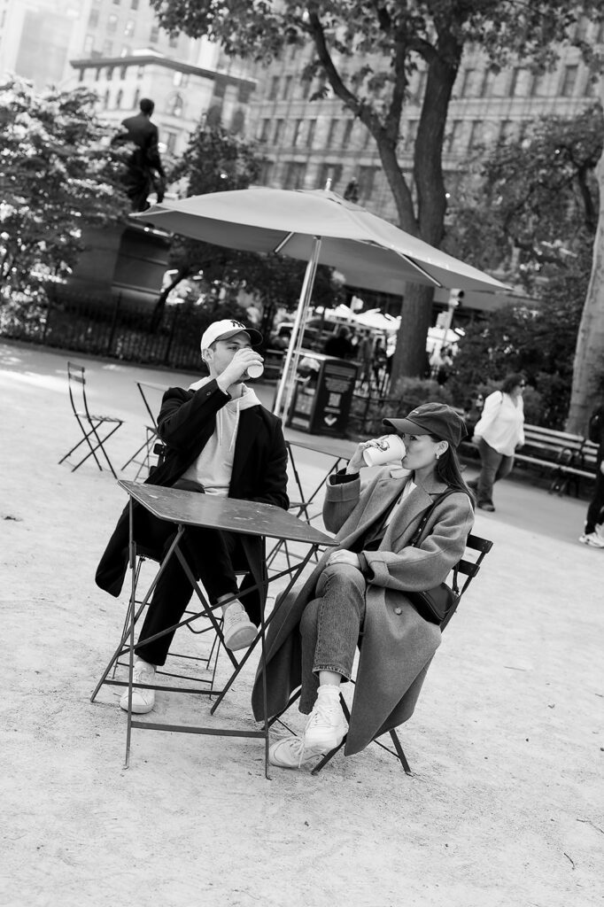 Black and white photo of a couple sitting at an outdoor café table sipping coffee during their Greenwich Village engagement photos.