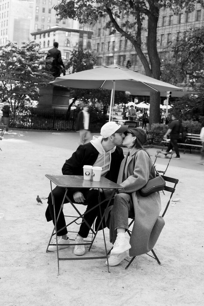 Black and white photo of a couple kissing across a café table with coffee cups in view during their engagement photos in NYC.