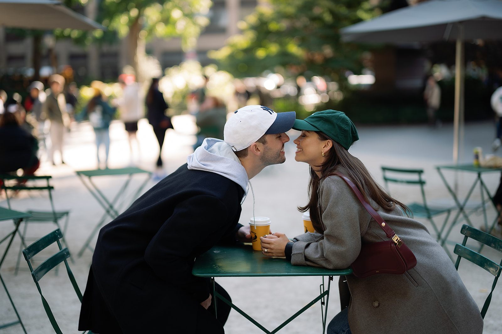 Color photo of a couple smiling at each other across a green café table while holding coffee cups during their cozy NYC engagement photos.