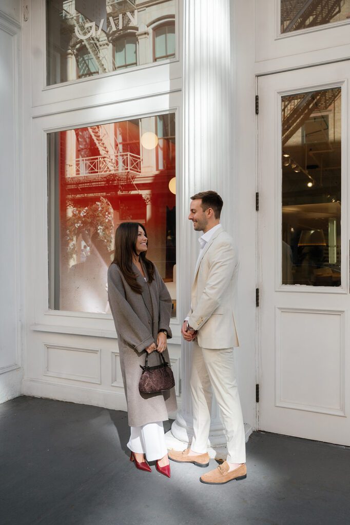 Couple smiling at each other in front of white storefront in SoHo during their NYC engagement session.