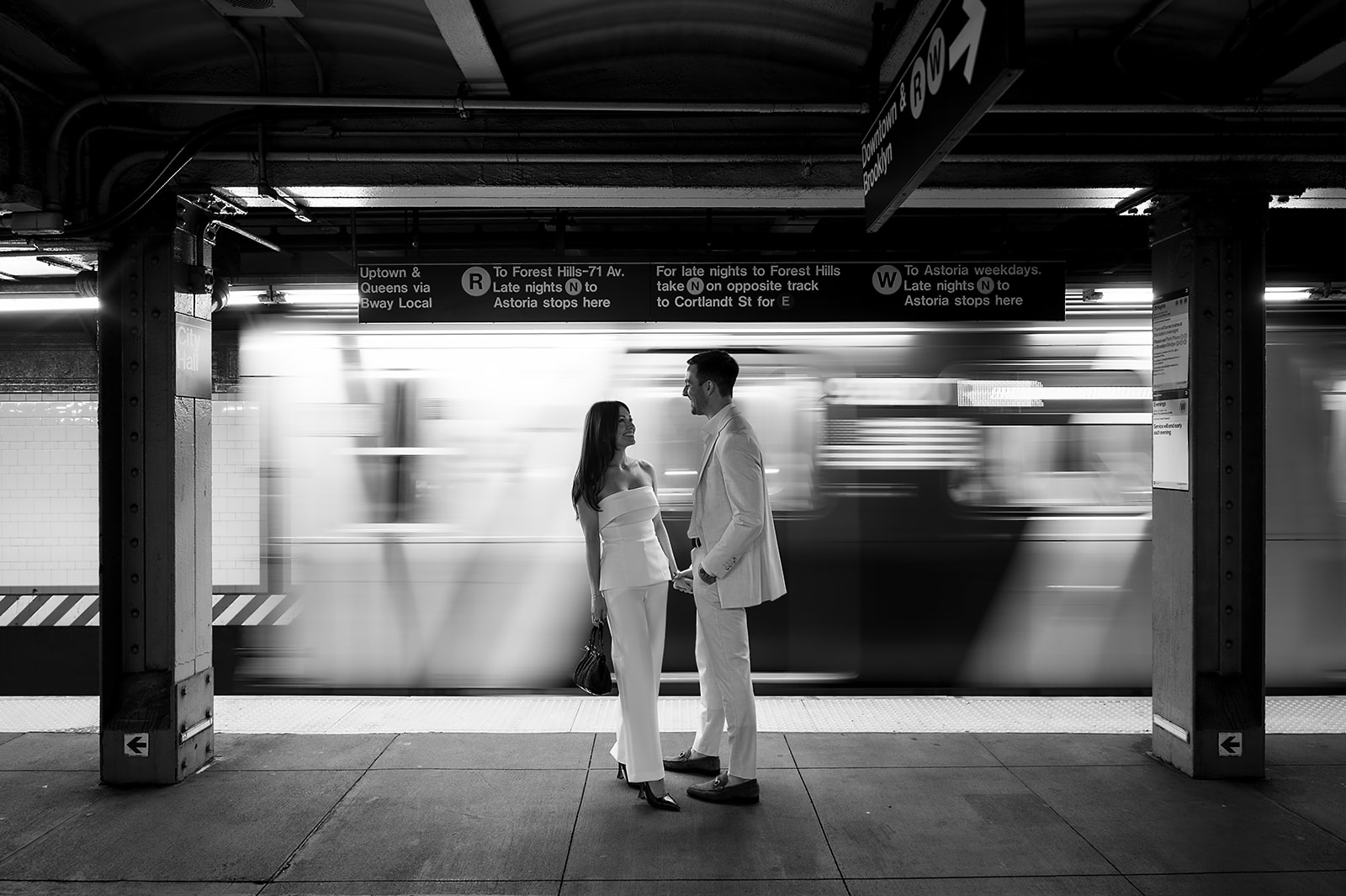 Couple holding hands on subway platform with motion blur of train in background during NYC engagement photos.
