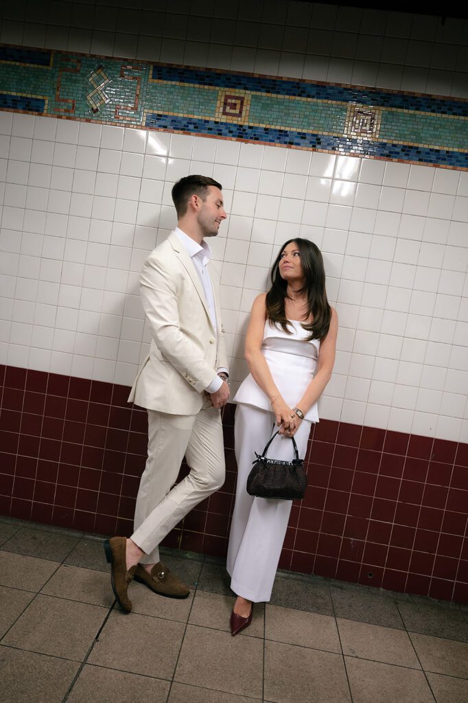 Couple standing against tiled subway wall looking at each other.