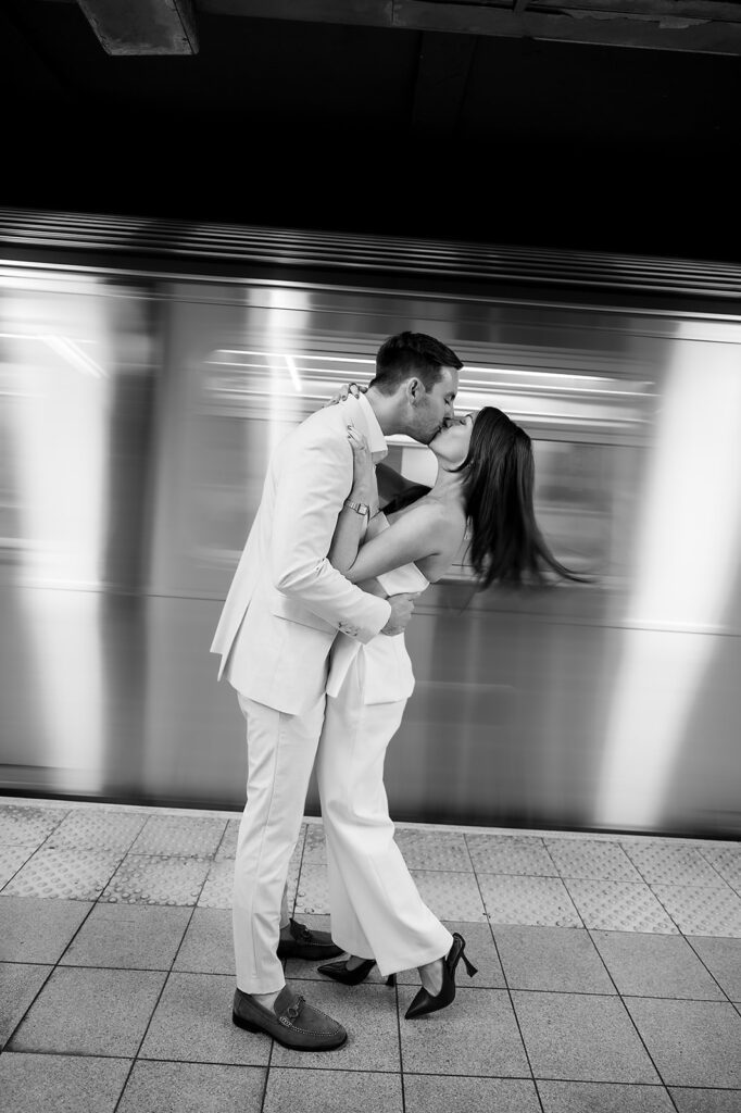 Black and white NYC engagement photo of couple kissing on subway platform with train passing.