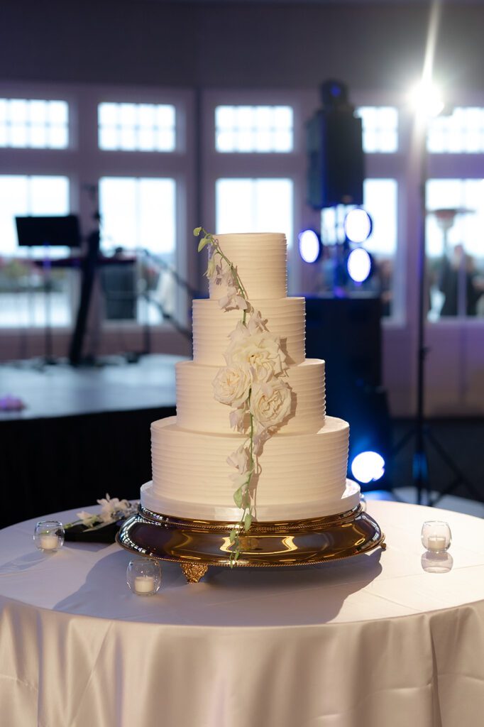 Four-tier white wedding cake with cascading white flowers on a gold stand inside the Lange Center Ballroom at Bay Harbor Yacht Club.
