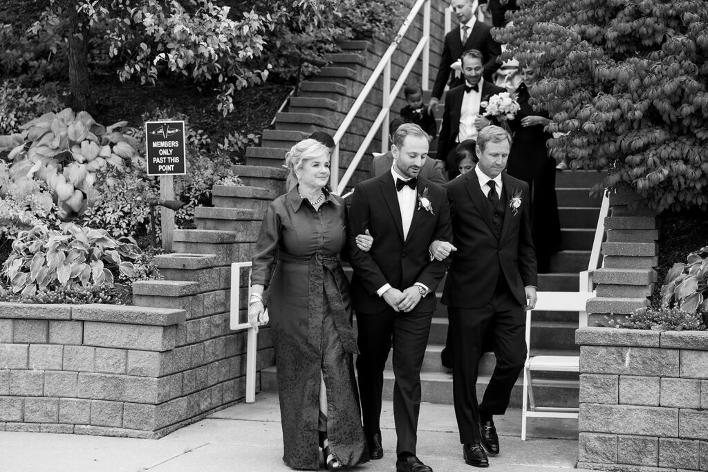 Groom walking arm in arm with his parents down the steps toward the ceremony.