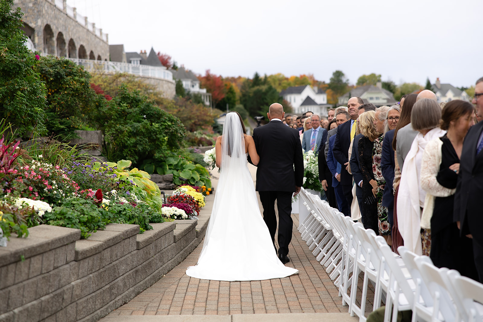 Bride walking down the aisle with her father surrounded by guests and colorful garden blooms.