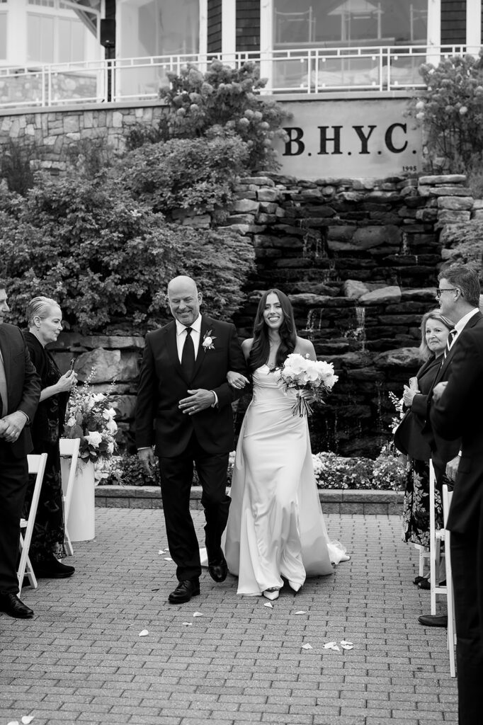 Bride smiling as she walks with her father toward the groom in front of the BHYC sign and waterfall.