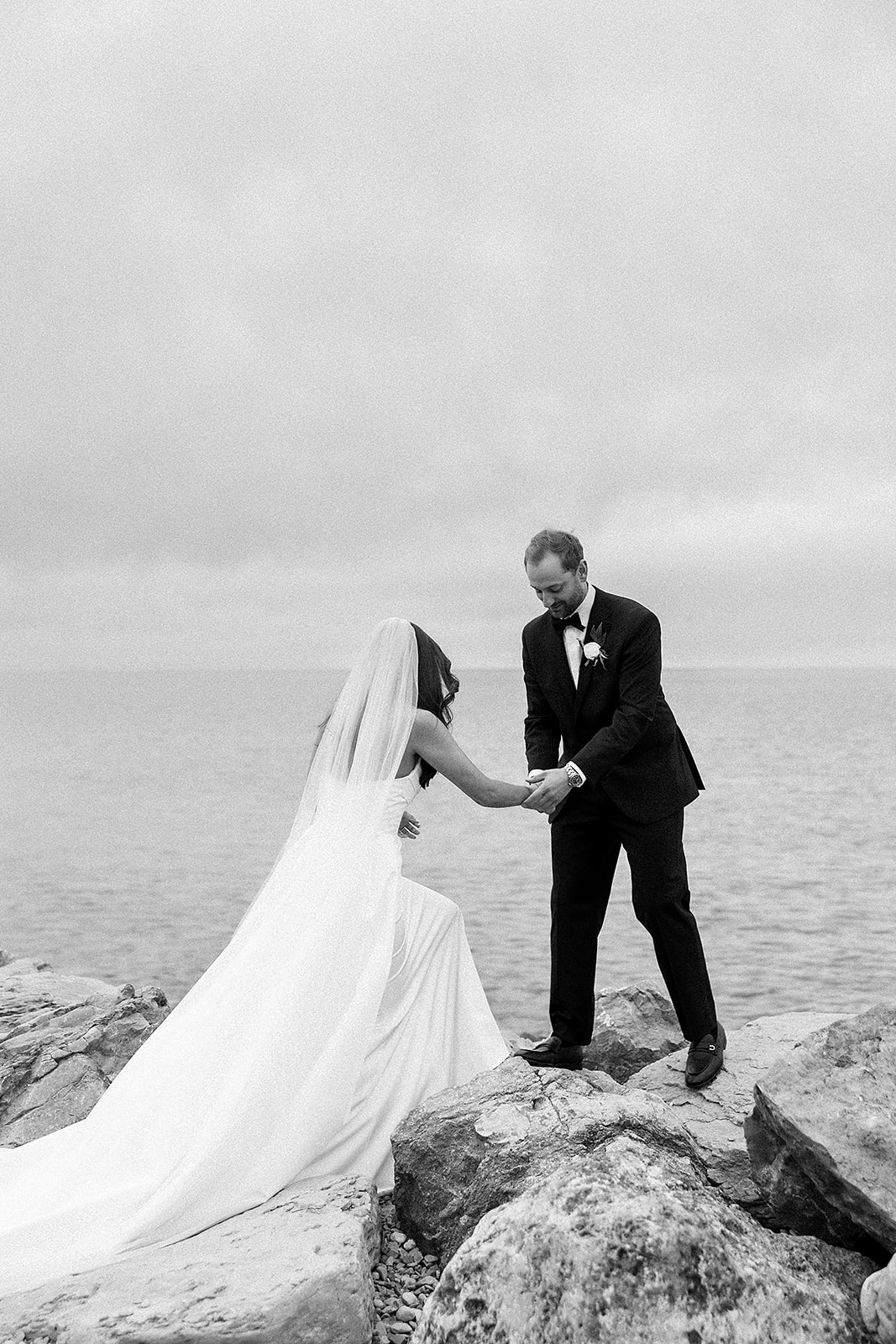 Groom helping the bride across rocks by the bay for portraits