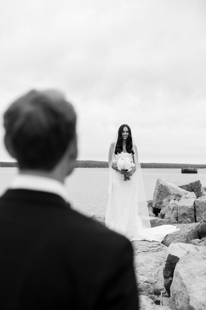 Bride standing on the rocks by the water smiling toward the groom.