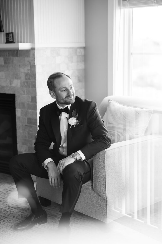 Groom sitting near a window in a black tuxedo and bow tie while getting ready.