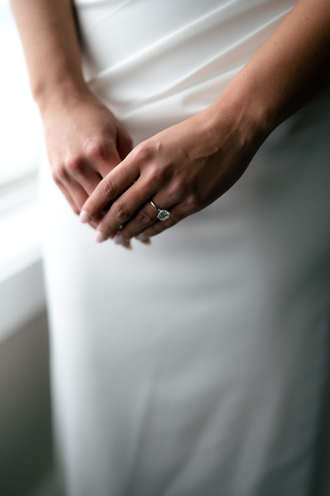 Bride’s hands resting together showing her diamond engagement ring against her satin gown.