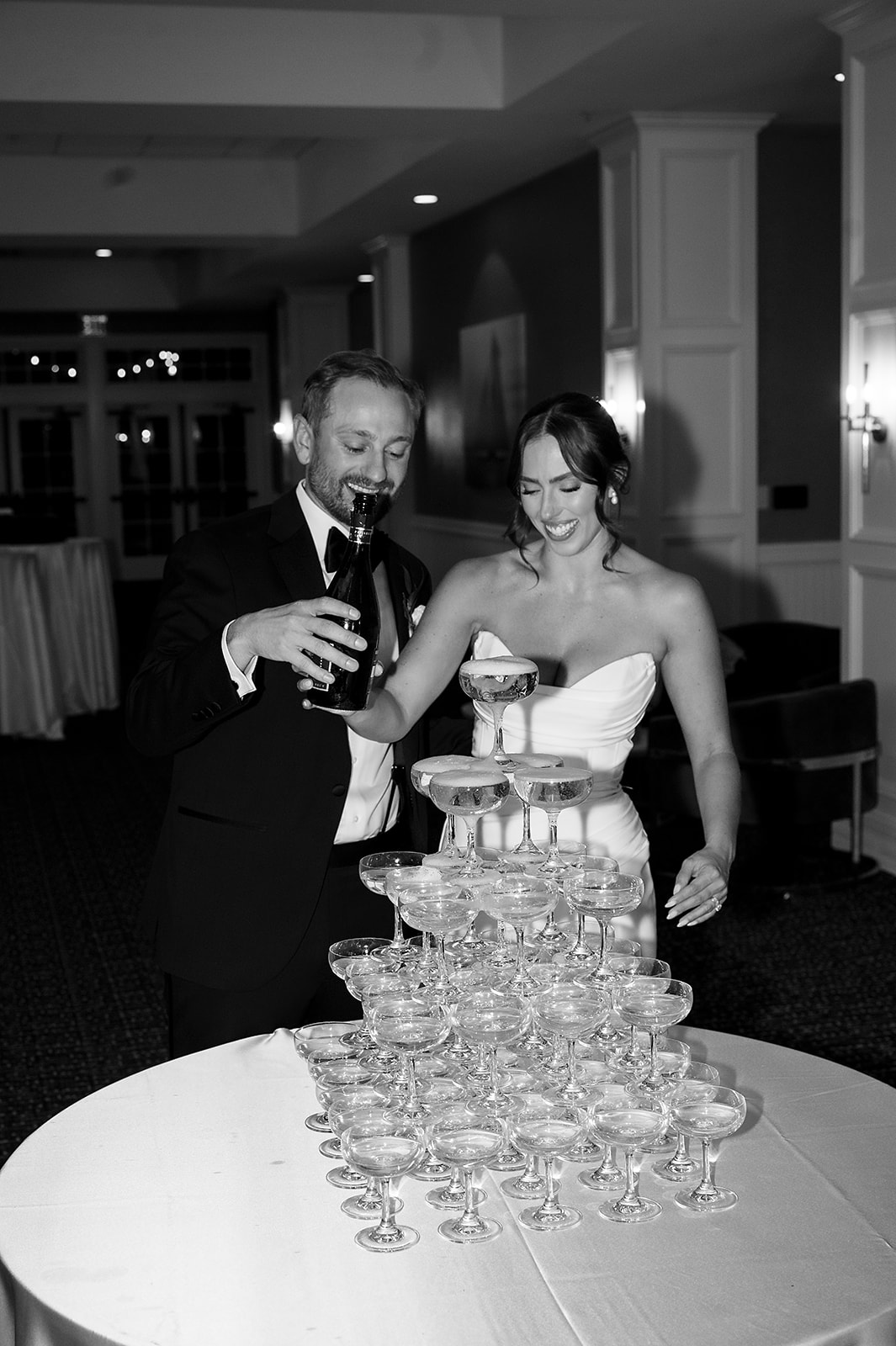 Black and white photo of a bride and groom pouring champagne into their champagne tower during their Bay Harbor Yacht Club wedding reception in Northern Michigan