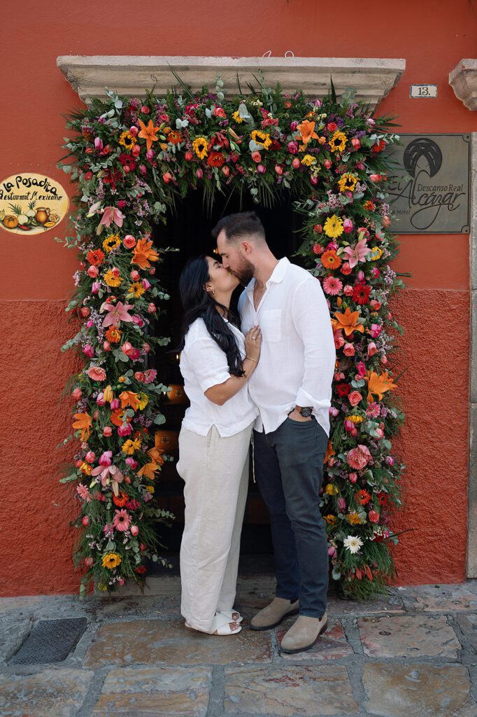 Couple sharing a kiss under a vibrant floral arch outside La Posadita restaurant.