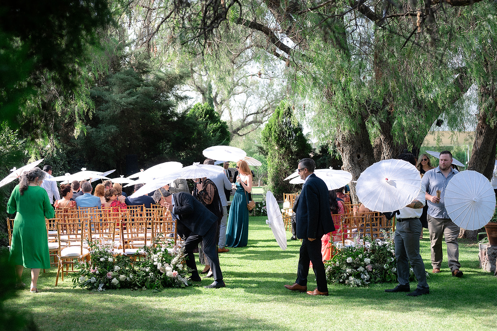 Wedding guests seated outdoors under trees holding white parasols