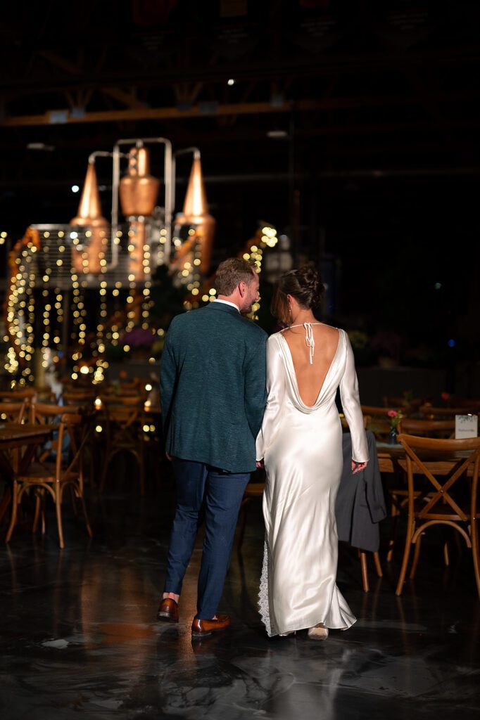 Couple walking hand-in-hand across the warmly lit Bay Harbor venue, her satin gown trailing behind as string lights glow in the background.