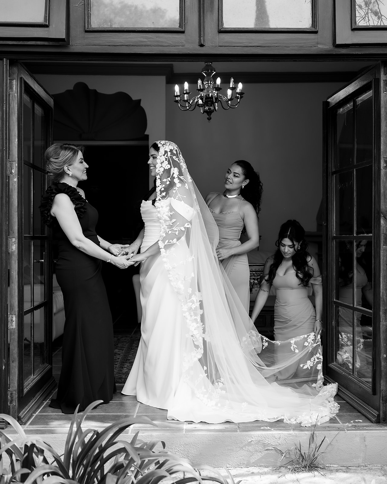 Black and white photo of a bride holding her mothers hands while the bridesmaids adjust her train for her San Miguel de Allende Wedding at Hacienda San Luis Gonzaga