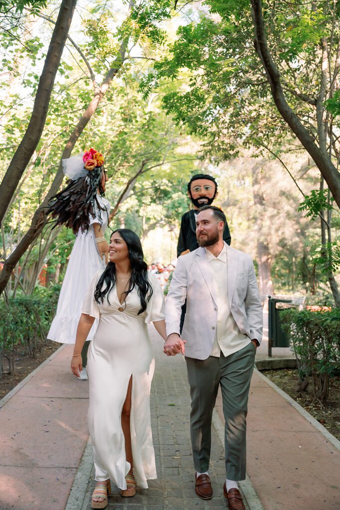 Bride and groom walk hand in hand during their San Miguel de Allende wedding callejoneada parade surrounded by traditional giant puppets and greenery.
