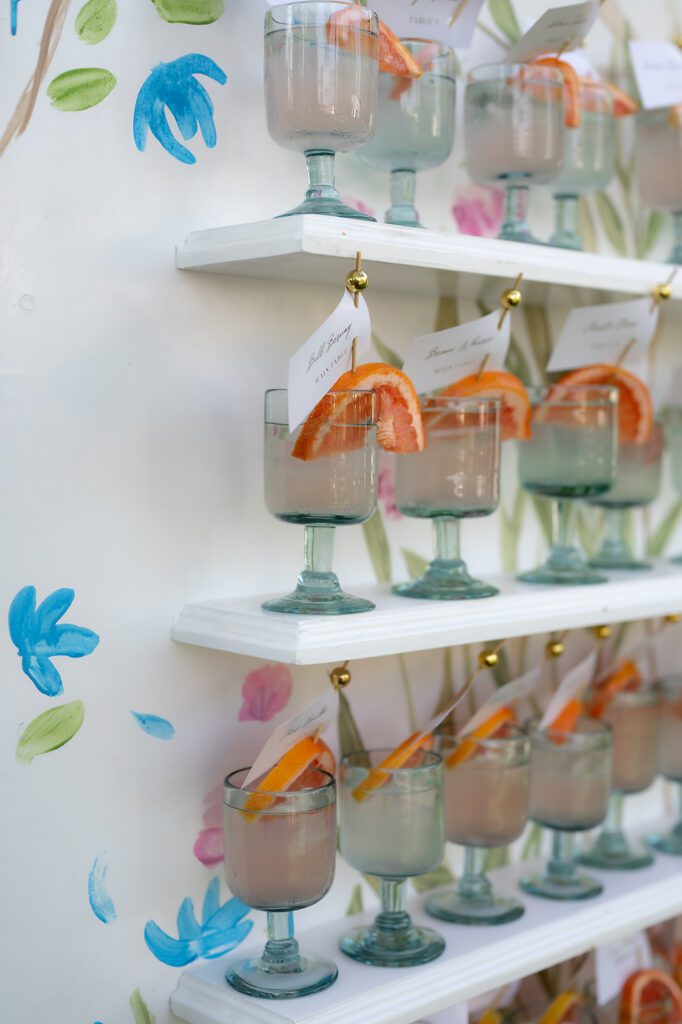 Colorful escort wall display at Hacienda San Luis Gonzaga wedding reception, featuring pastel floral artwork and glass goblets filled with grapefruit cocktails.