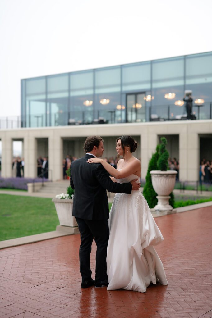 Bride and groom sharing their first dance outdoors while guests watch from the patio at Grosse Pointe War Memorial during cocktail hour.