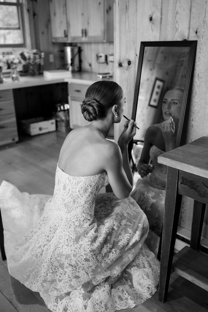 Bride applying lipstick in a mirror while wearing her lace wedding dress.