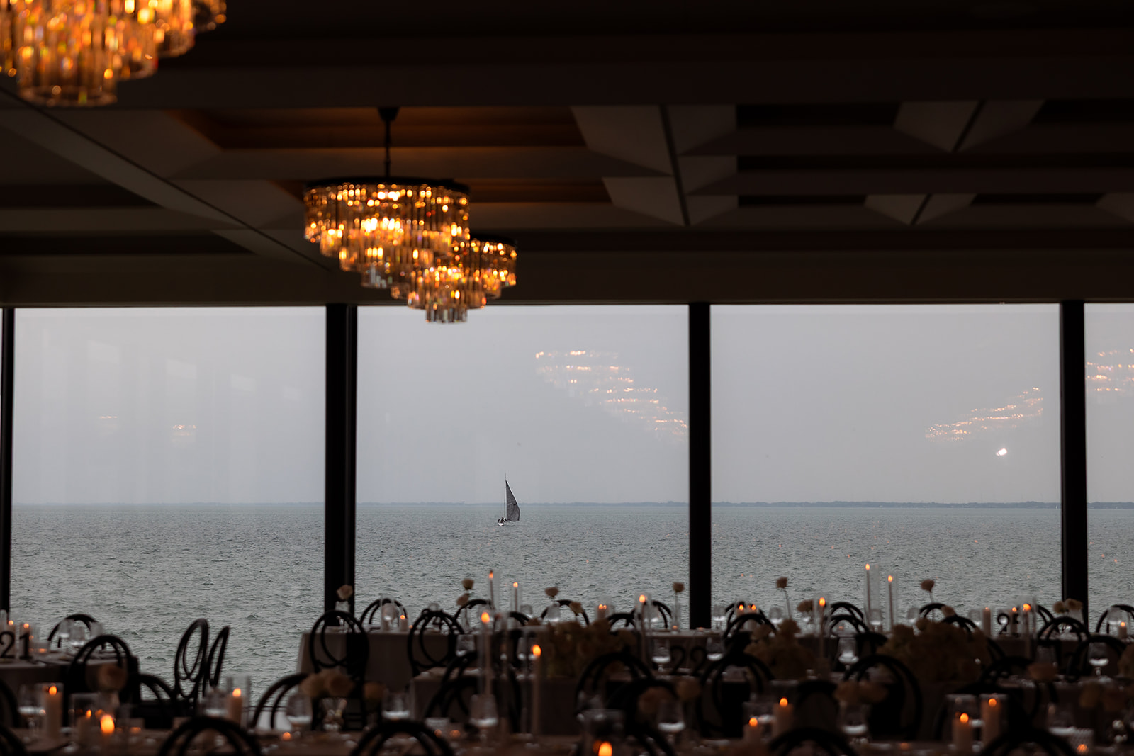 Elegant ballroom reception setup at Grosse Pointe War Memorial with candlelit tables, black chairs, and panoramic lake views through floor-to-ceiling windows.