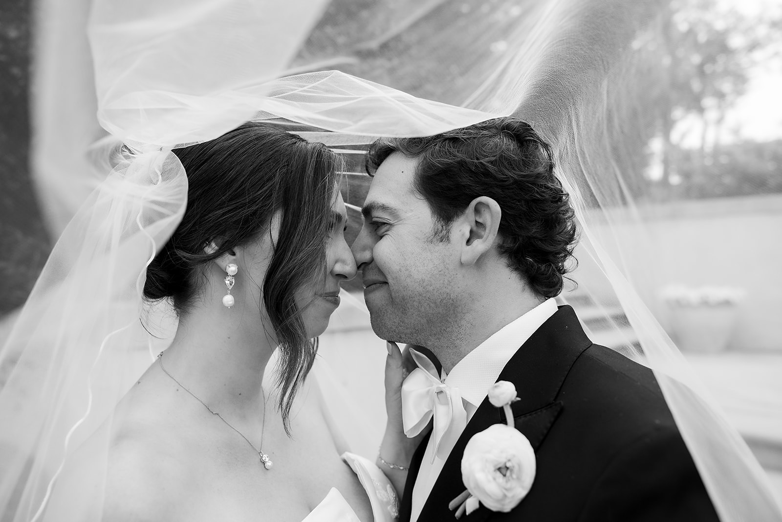 Black and white wide shot of a bride and groom posing underneath a veil. 
