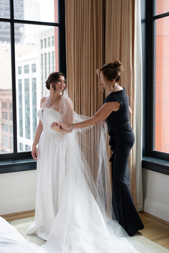 Bride getting ready in her wedding dress with help from her mother near the window at the Shinola Hotel.