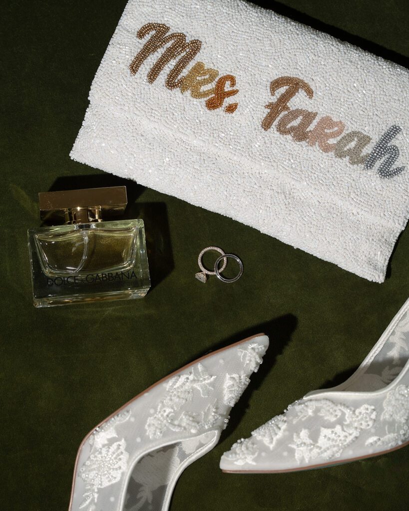 Wedding details flat lay with bride’s shoes, rings, perfume, and personalized clutch at Shinola Hotel.