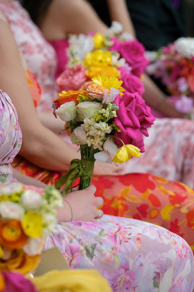 Close-up of bright bridesmaid bouquets resting on floral dresses during an outdoor Northern Michigan wedding ceremony.