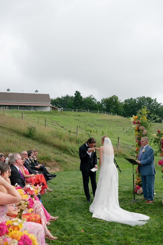Bride and groom stand at the ceremony altar at Noverr Farms as the groom kisses the bride’s hand during vows.