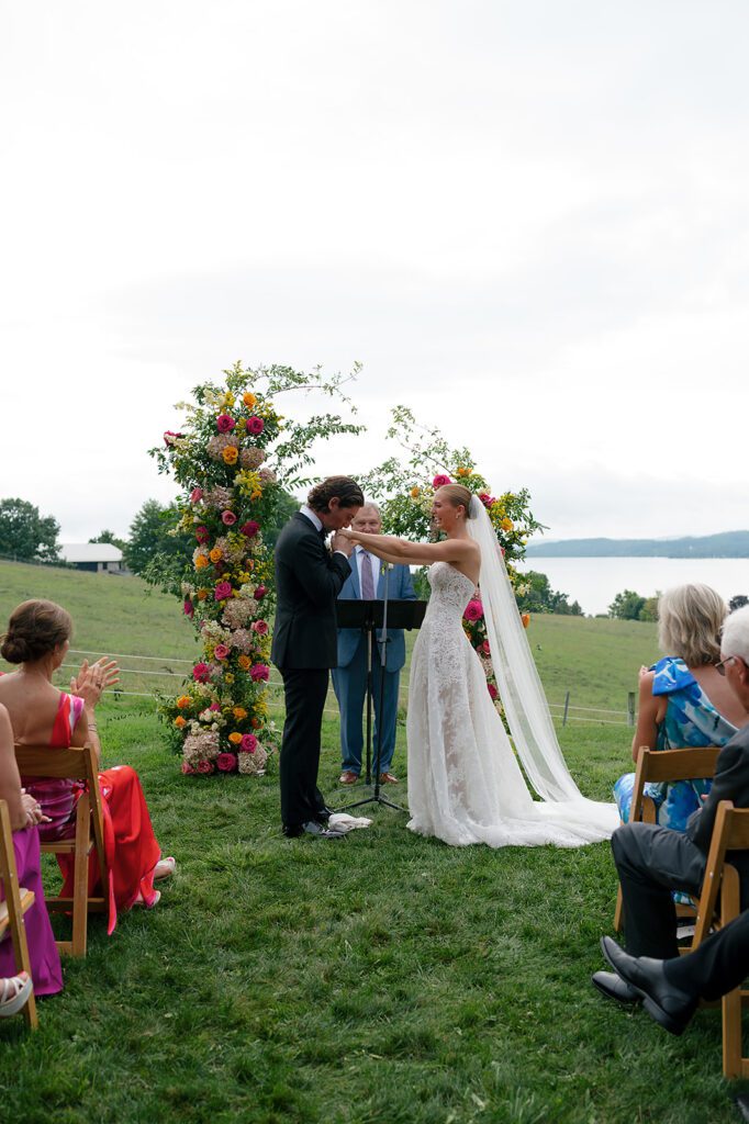 Wide view of the couple exchanging vows beneath floral pillars at a Noverr Farms wedding with Lake Leelanau behind them.